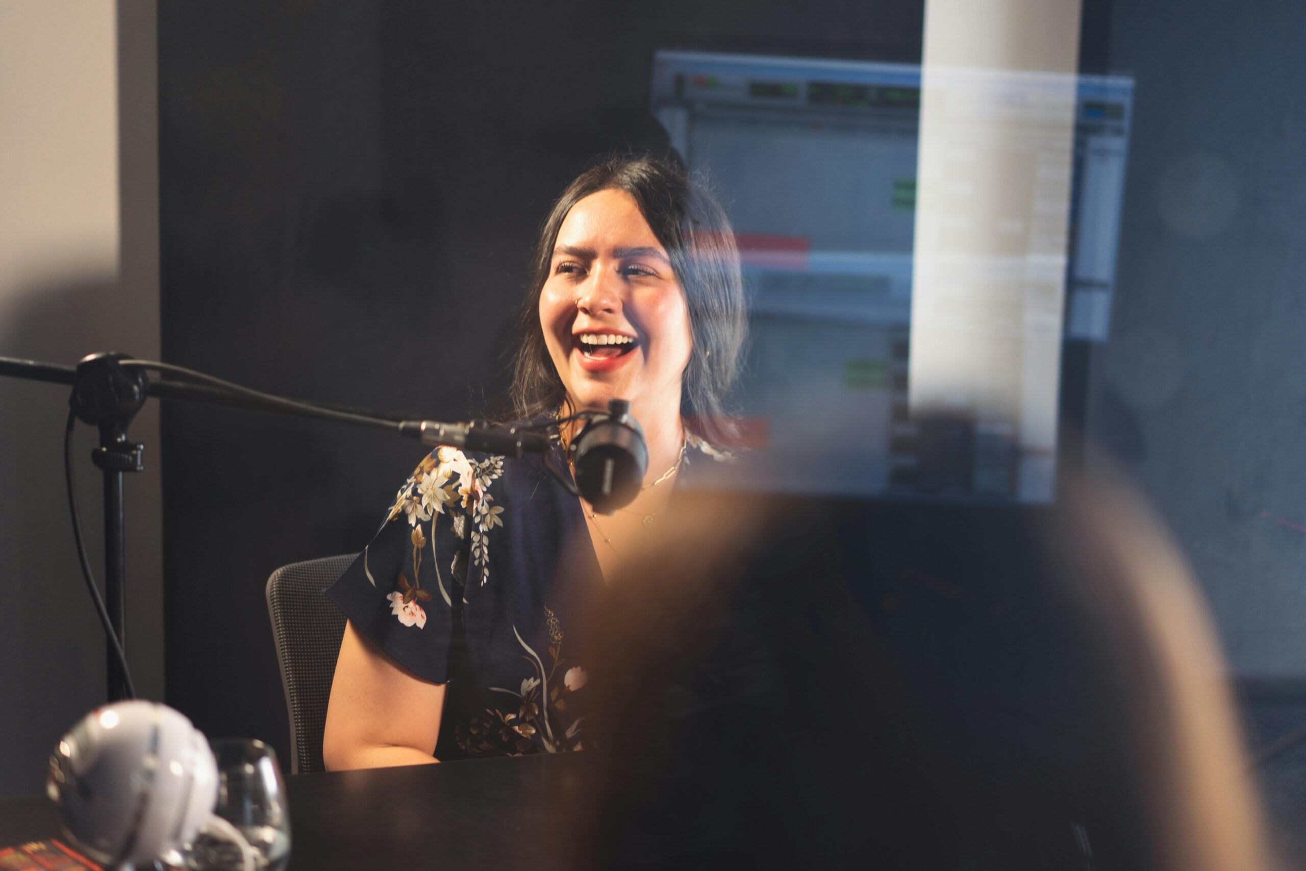 A woman smiles while recording a podcast indoors with a microphone.